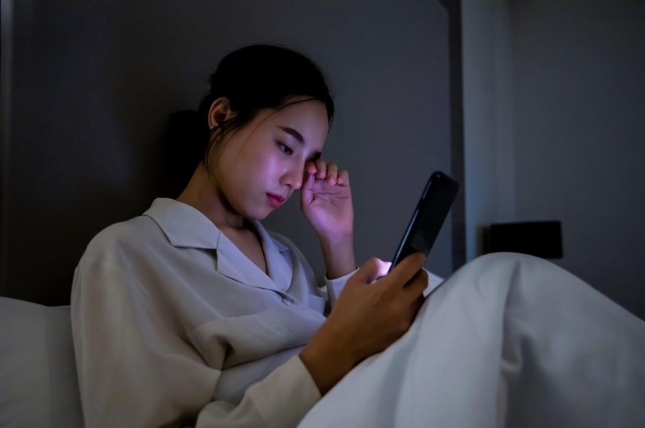 Young woman feeling tired, rubbing her eye with one hand and holding her phone in the other in a dimly lit bedroom.