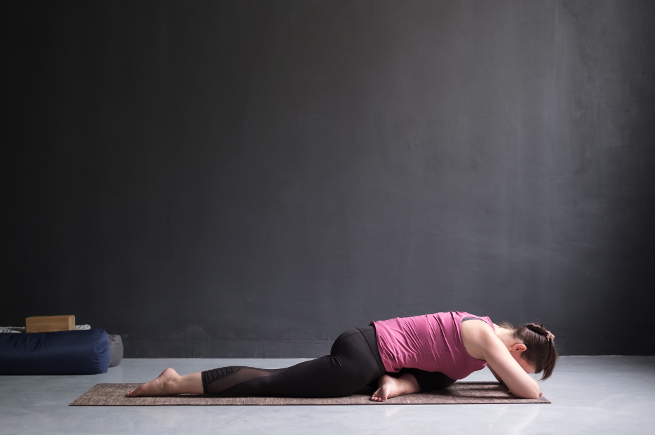 Woman in pink vest and black leggings in a lying pigeon yoga pose to release stress from the body and release tension from the hips.