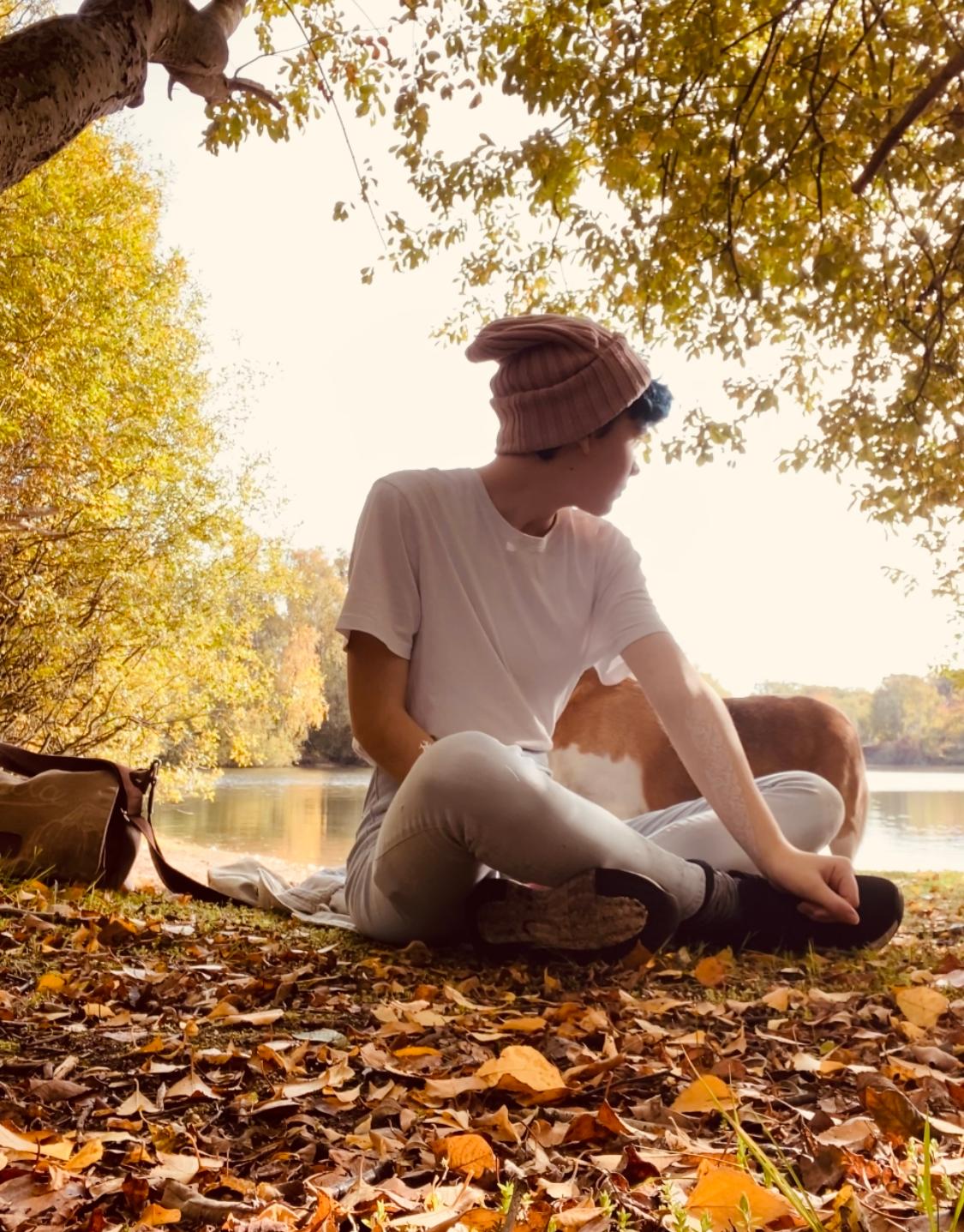 Teen sat outside in nature by a lake during September in the autumn leaves, under a tree wearing a bobble hat. Teen stress. Back to school stressors.